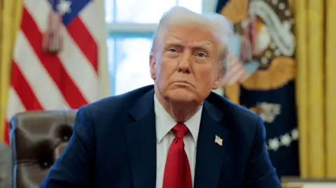 Getty Images Donald Trump lleva una chaqueta de traje azul marino con un alfiler de la bandera estadounidense, una camisa con cuello blanco y una corbata roja. Está mirando hacia arriba con el ceño fruncido mientras se sienta en la Oficina Oval, que tiene dos banderas a ambos lados de él.