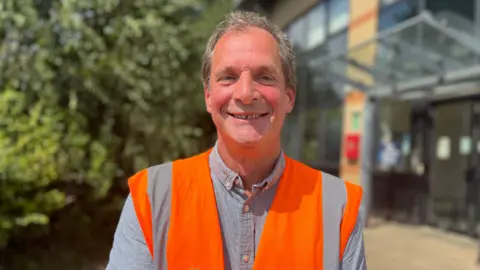 Nicola Haseler/BBC A man with grey hair, a grey shirt and a bright orange high-vis jacket stands smiling in front of the Buntingford waste depot. 