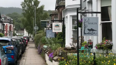 A general view of a street in Keswick lined with holiday accommodation signs, outside terraced houses with bay windows. Cars are parked all along the street.
