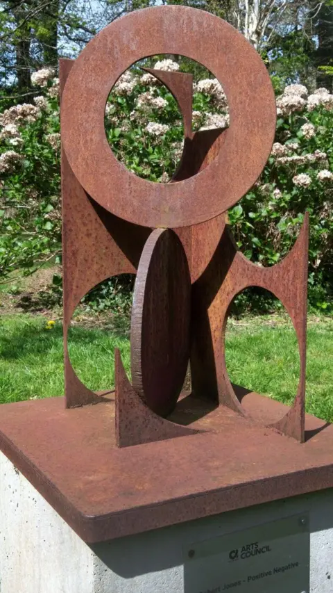 ROBERT JONES A metal sculpture featuring interlocked circles. It is on a concrete plinth in the arboretum.
