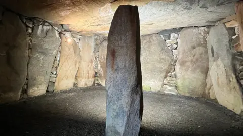 A central horizontal stone is inside a stone circle with large stones around it and large pieces of stone forming a roof.