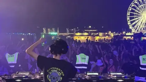 A young boy wearing a black T-shirt saying "DJ Hypester" on it and a pair of black headphones, DJing in front of a crowd of people at night. He's got his back to the camera and is facing the crowd, who have their arms in the air. Behind them is a Ferris wheel and some lit-up festival food stalls.