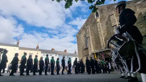 St John Ambulance volunteers line up to enter St James Church in Torpoint, with men with drums standing to the left. 