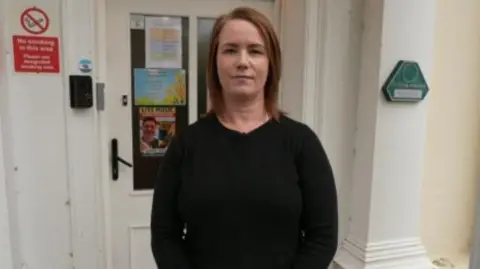 A woman with shoulder length auburn hair stood outside a building. She is wearing a black jumper.