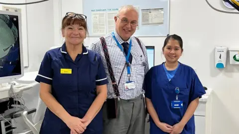 Northampton General Hospital Two women in blue medical scrubs stand either side of a man in glasses, a blue and white shirt with a tucked in tie and maroon suspenders and grey trousers. They're standing in a screening room.