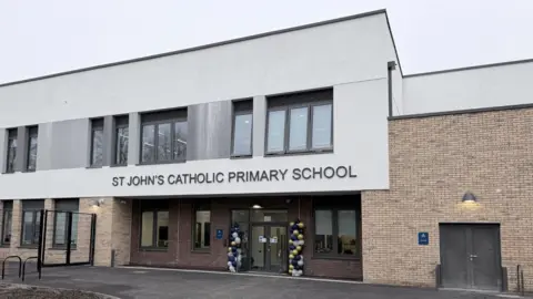 A newly built two‑storey school building with a modern design. The entrance features glass doors decorated with blue and yellow balloons, and signage above reads “ST JOHN’S CATHOLIC PRIMARY SCHOOL”. The exterior combines light brick and white cladding, with large windows across the upper floor.