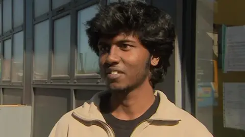 Loges standing outside the flats is wearing a brown jacket. He has a thin beard and black hair. 