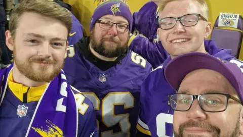 Jonathan Fry Four people including Jonathan (top right) are wearing American football jerseys and sitting watching a game. They are all smiling and looking at the camera.