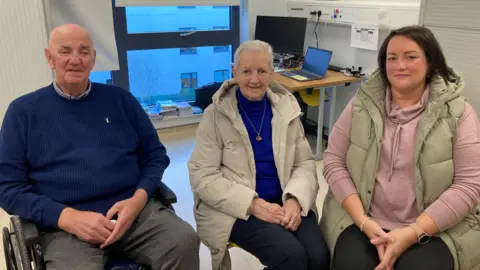 Jim Ross wearing a white and blue checked shirt under a blue jumper. His wife Lorna wearing a blue jumper, a beige waterproof hooded coat and a gold necklace. His daughter Gayle wearing a pink hoodie with a beige hood bodywarmer. All three are sat down in an office with a window and a laptop beside a monitor on a wooden desk behind them. 