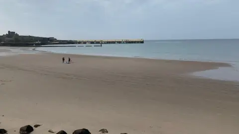 A sandy beach at low tide. There are two people on the beach with a dog. Peel Castle is to the left in the distance.