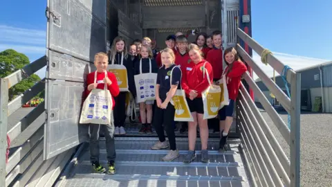 A group of primary school children wearing red and navy jerseys stand at the back entrance to an HGV transporter