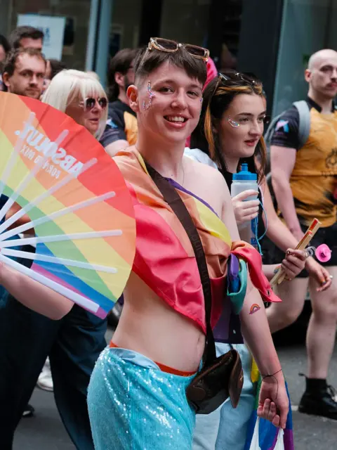 A Pride participant wearing a colourful flag and a blue sequined skirt holds an open rainbow folding fan while walking in a parade.