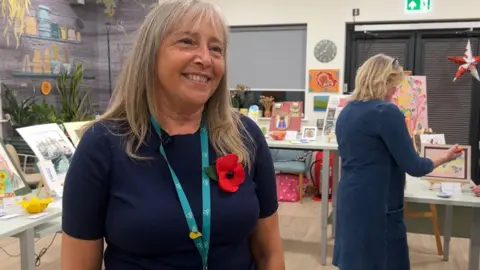 Rebecca Richardson stands in a room full of artworks, some are hanging on the walls, some are in frames on tables. Rebecca is a white woman who has long grey hair with a fringe and is smiling. She is wearing a blue woollen jumper with short sleeves and a brooch of a large red poppy flower. She also wears a long blue lanyard ribbon with the logo of the Severn Hospice, which is in a love heart shape. Around her, a woman with blonde hair and a blue dress with long sleeves examines some of the art work. 