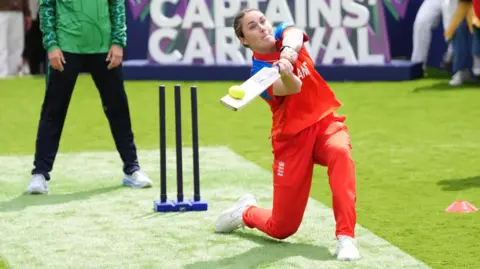 England captain Nat Sciver-Brunt, in England's red and blue kit, swings a bat to hit a ball during a game in front of a 'Captains' Carnival' sign, watched by Ireland captain Gaby Lewis in green