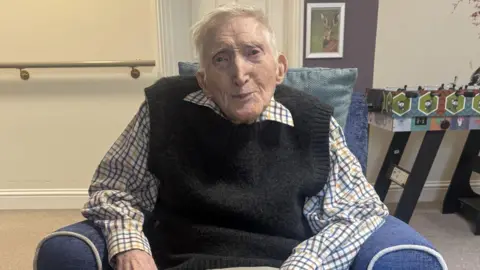 An elderly man sits in a blue arm chair and smiles at the camera. He has short grey hair and wears a shirt with a black sleeveless jumper over the top. 