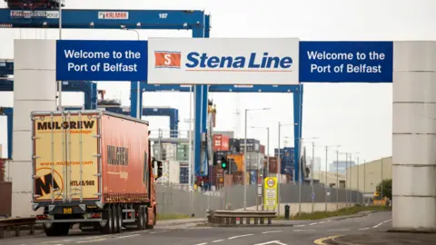 Image of freight lorry driving through entrance of Belfast port. Blue signage above the orange and yellow lorry reads 'Welcome to the Port of Belfast' on both ends and 'Stena Line' in the middle. 