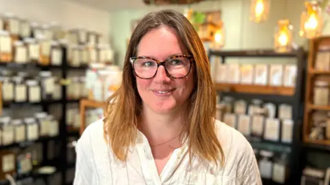 Steph Van de Pette has shoulder-length auburn hair and tortoiseshell glasses. The shelves of her shop behind her are filled with large storage bottles.