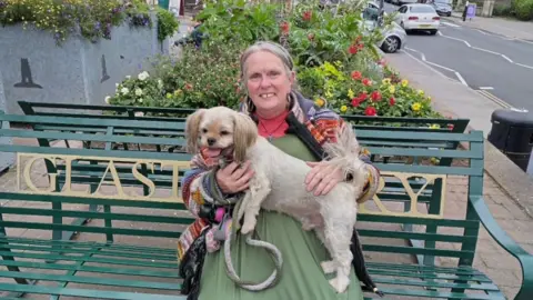 Women with a dog sat on a green bench that says Glastonbury. She has grey hair and is wearing a green dress. There's a flower bed in the background.