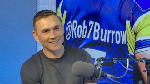 Kevin Sinfield sat in a blue studio, smiling