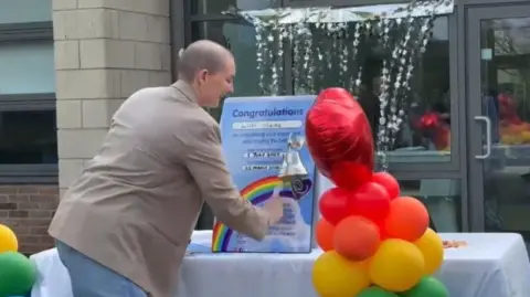 Handout A woman with a bald head is wearing a beige jacket and jeans and is smiling as she rings a silver bell on a white table. A board on the table says congratulations Lilli Hales and there is a colourful balloon display at either side of the table with a red heart on the top.