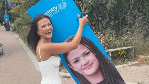 A woman with brown hair and a white dress holding a blue cardboard cutout showing her face.