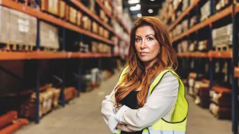 Woman in a warehouse with red hair and a hi-vis jacket.