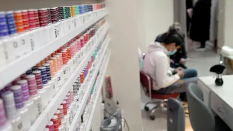 A nail technician in a nail bar, with rows of nail varnish products in the foreground