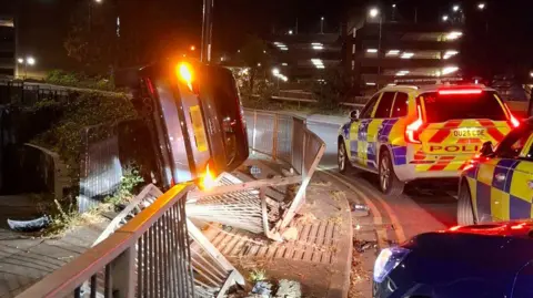 Two police car on the road with a car on its side on the roundabout - inside the metal barriers. 