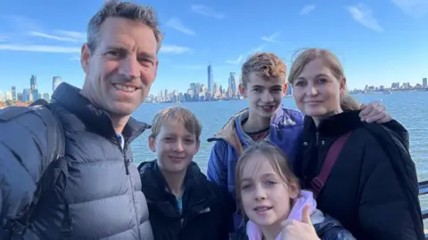 Charlie, on the left, is stood with his children, Harry, Tom and wife Mel, with their daughter Rosie in front of them, in front of a body of water, all smiling with buildings in Manhattan, New York, in the background. 