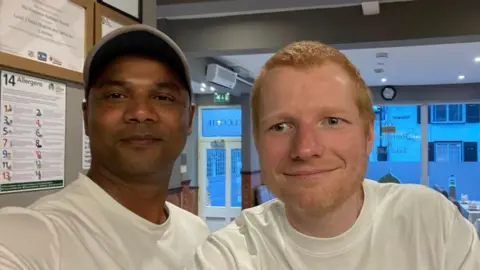 Mitu Belan A man wearing a grey cap and white T-shirt takes a selfie. A man with red hair and a long sleeved white T-shirt stands next to him. They are both smiling at the camera. They are standing in the front part of a curry house. 