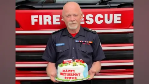 A bald man standing in front of a red fire engine. He is in a navy firefighter uniform and is holding a cake that is white and blue and has two red fire engines on the top of it. red lettering on the cake says "happy retirement steve"