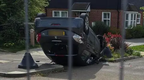 A black car flipped over on its roof, with a house in the background.