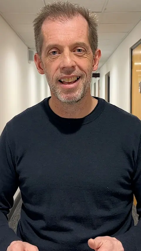 A white man with brown hair in a black shirt looking at the audience.