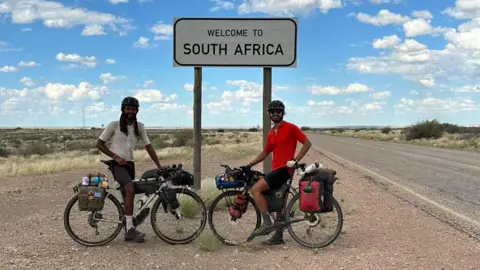 Contributed Two men sitting on bikes next to a deserted road. They rest in front of a sign that reads Welcome to South Africa. They both wear bike helmets and one wears a white cycling top and the other wears a red one. 