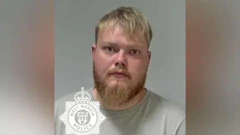 A police mugshot of a man with blonde hair and a beard. He is wearing a grey T-shirt.