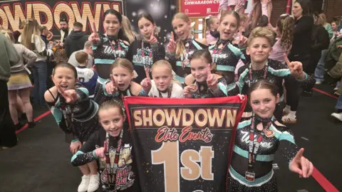 Ten young people gathered around a 1st placed sign. They are wearing leotards and holding up the a number one finer on their hands They are at a competition with lots of people milling around behind.