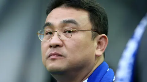 Dejphon Chansiri looks at the camera. He has short, dark hair wire-framed glasses and is wearing a blue and white scarf.