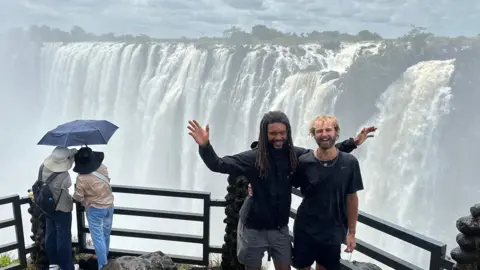 Contributed Two men stand together on a ledge that overlooks a large waterfall. One has their arms outstretched in celebration.