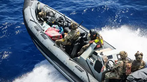 Royal Navy A rigid inflatable boat containing four personnel in camouflage outfits, seen from above. The boat appears to be cresting a wave in a choppy sea. 