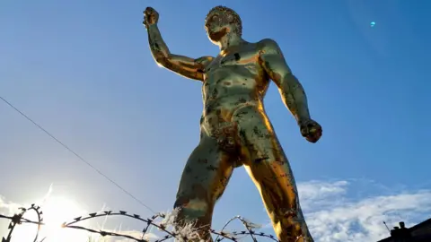 The BBC's front cover from below is a large golden statement of a male gladiator with short hair on his head and a fan raised in the air. It is naked and male and has small appendages. The blue sky and rooftops can be seen behind the big statue. 