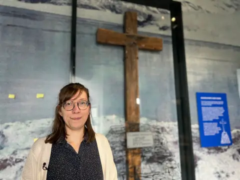 A woman with a black and white dress and a cream cardigan stands in front of a large wooden cross that has been fixed to a wall.