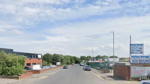 A road in an industrial area of Leeds with a garage building on one side and a red brick building on another. A few cars are parked on the street.