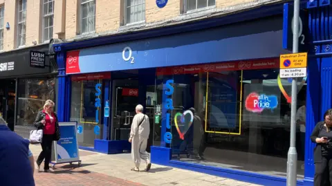 View from across the road of an O2 phone shop. The outside is blue and has sale signs either sides of the doors. People are walking past the shop. 