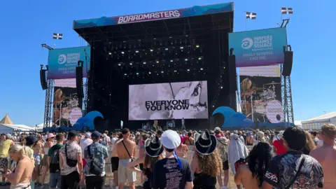 A crowd of people stood in front of the Boardmasters stage watching a live performance. It is a sunny day with clear blue skies, and many people are wearing hats. The stage has blue and purple ombre signs around it, and the screen at the centre of the stage says 'Everyone you know'.
