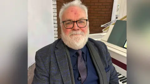 BBC Alan Gregory, piano teacher, sits at a white piano in a dark three piece grey and navy suit. He has grey hair, a grey beard, blue eyes and wears glasses. 