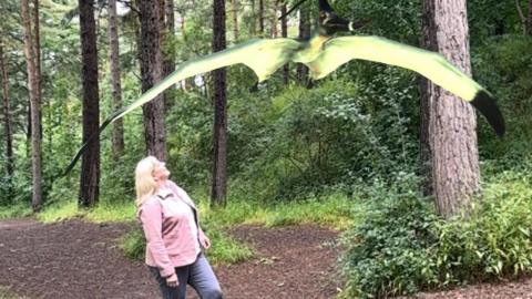 Helen Howell with long blond hair and a pink top looks up at a pterodactyl in a wood 