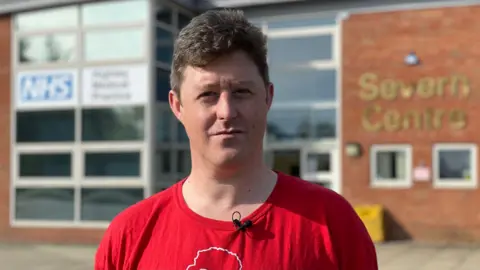 Man wearing a red t-shirt posing in front of a red-bricked community building with white-framed windows and an 'NHS' sign above the entrance