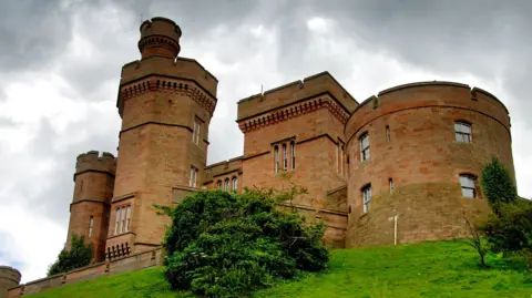 A view up a grassy embankment to some of the towers of sandstone-built Inverness Castle. The sky above is grey and cloudy.