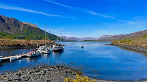 A body of water surrounded by hills with boats moored off the jetty. The sky is clear and the water is bright blue.