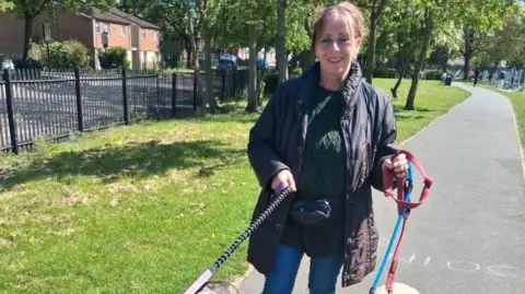 Clair Igoe A woman with dark hair that is tied back is smiling. She holds a dog lead in each hand. she wears a dark coloured puffa jacket and is wearing a bumbag. 